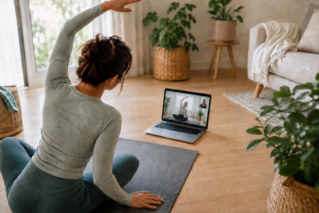 A woman stretching on a mat at home while following a live coaching session on her laptop.